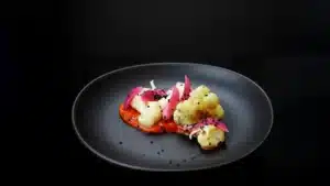 "Plato gourmet de coliflor asada acompañada de cebolla morada en vinagre, sobre una salsa roja vibrante, decorado con semillas de sésamo negro, presentado en un plato oscuro con una iluminación suave que resalta sus texturas y colores."