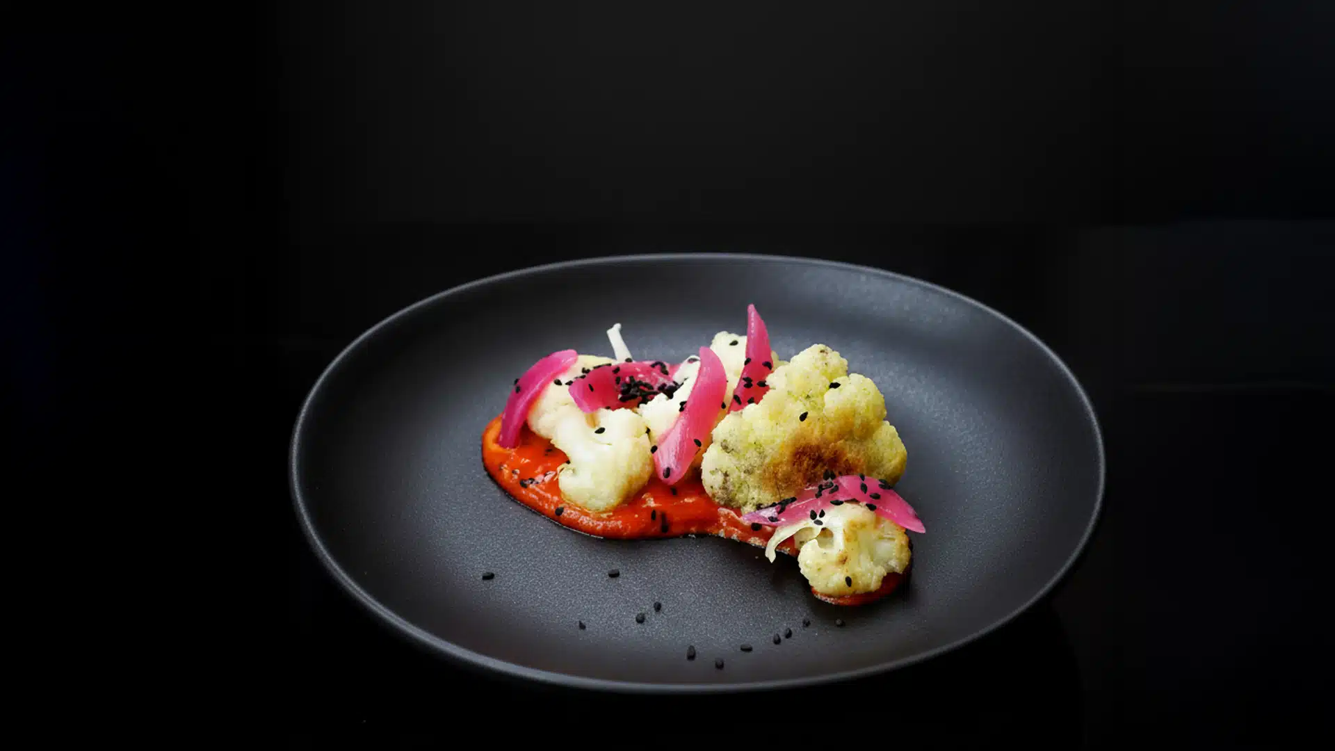 "Plato gourmet de coliflor asada acompañada de cebolla morada en vinagre, sobre una salsa roja vibrante, decorado con semillas de sésamo negro, presentado en un plato oscuro con una iluminación suave que resalta sus texturas y colores."